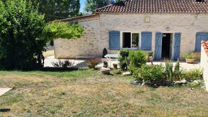 Une maison en pierre avec un toit de tuiles, des volets bleus et un jardin agrémenté de plantes en pot et d’une petite table.