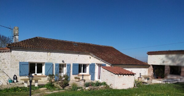 A rustic stone house with a tiled roof, blue shutters, and a small garden area with a bench and lamp post. - Country house with swimming pool (Beauville)