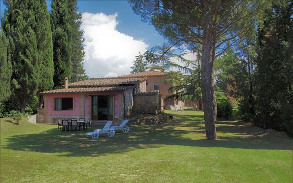 Uma casa rústica com telhado de telhas, um pátio com cadeiras e uma mesa, e um gramado.
