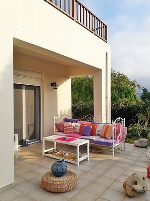 A covered outdoor seating area with a sofa and a table, a balcony with a railing, and a view of greenery. - Villa Armonia, Everything Is In Harmony (Agios Nikolaos)