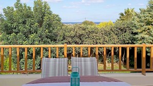 Un espace extérieur ensoleillé, aménagé avec des sièges, une table pour deux, une rambarde en bois et une vue sur un jardin luxuriant et un paysage lointain.