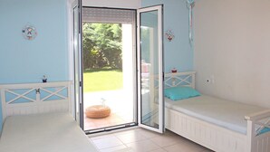 Une chambre avec deux lits blancs, dont l’un est agrémenté d’un oreiller bleu, et une grande porte-fenêtre coulissante donnant sur un jardin.