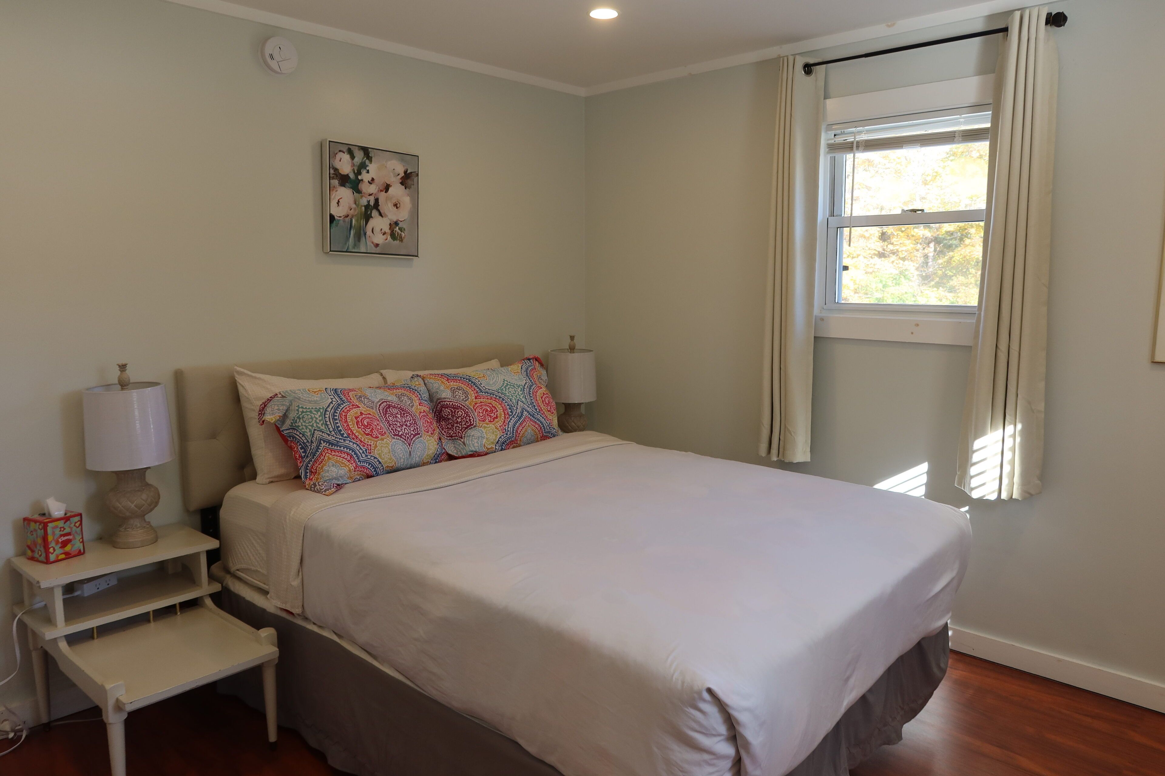 A bedroom with a bed, bedside lamps, a nightstand, a small table, and a window with curtains.