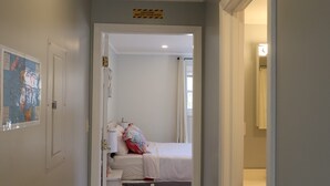 Un couloir étroit mène à une chambre à coucher qui contient un lit, une table de chevet et une commode. Une porte se trouve sur la gauche et on aperçoit une salle de bain sur la droite.