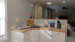 Une cuisine de taille réduite, équipée d’un évier, d’un plan de travail et de placards. On y trouve une fenêtre avec des stores, un ventilateur de plafond et un tableau accroché au mur.
