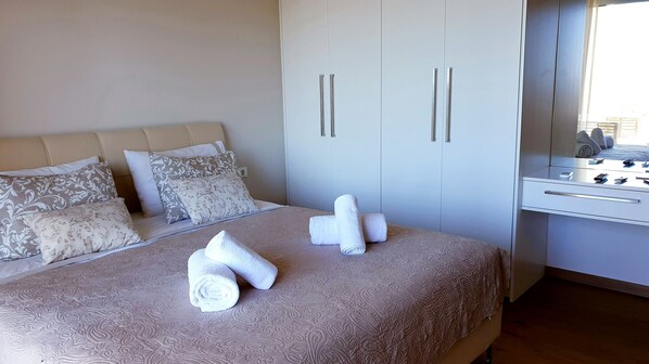 A neatly made bed with rolled white towels, a large wardrobe with two doors, and a small desk with a mirror and some items on it.