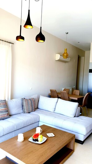 A modern living room with a light grey sofa, patterned cushions, a wooden coffee table with fruits and a cup, and pendant lighting.