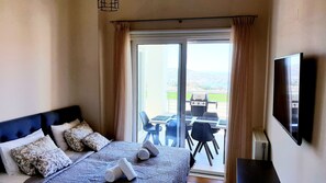 A hotel room with a bed, a TV, a balcony with a table and chairs, and a view of the outside.