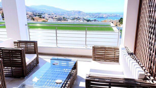A balcony with wicker furniture, a glass table, and a view of a coastal town and mountains.