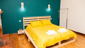 Uma cama com estrutura de madeira e roupa de cama amarelo vibrante, uma pequena mesa de cabeceira com um controle remoto e um aplique de parede.