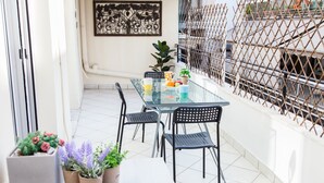 Mesa de vidro com cadeiras, plantas em vasos e um objeto decorativo numa varanda com soalho de azulejos brancos e vedação de metal.
