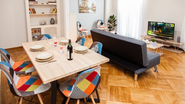 Uma sala de estar moderna com uma mesa de jantar posta para quatro pessoas, um sofá, uma estante e uma televisão.