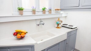 Uma cozinha com bancada branca, pia e armários. Há plantas em vasos no parapeito da janela e uma fruteira sobre a bancada.