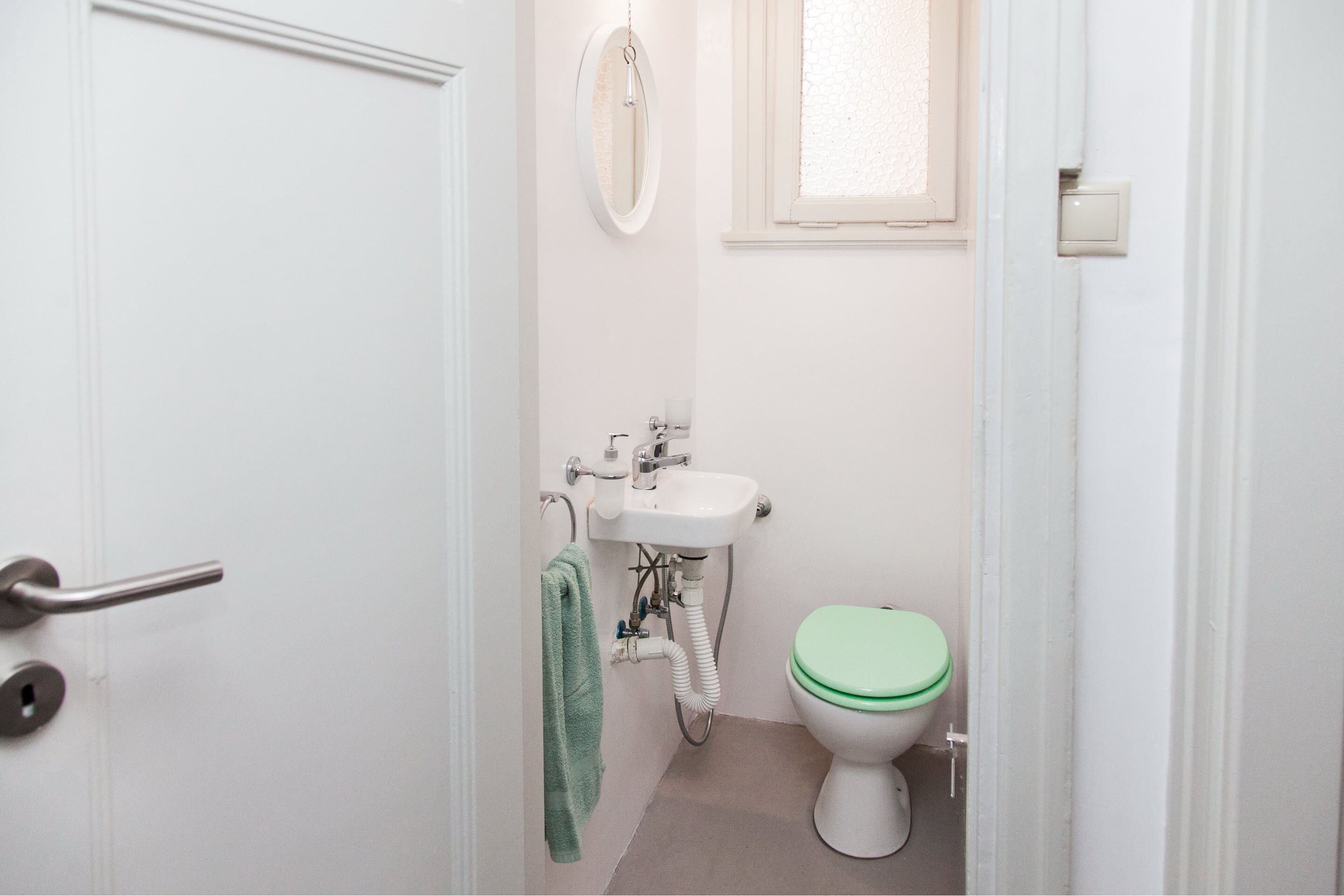 Un bagno piccolo e pulito con un lavandino bianco, uno sciacquone con il sedile verde e un asciugamano appeso al muro.