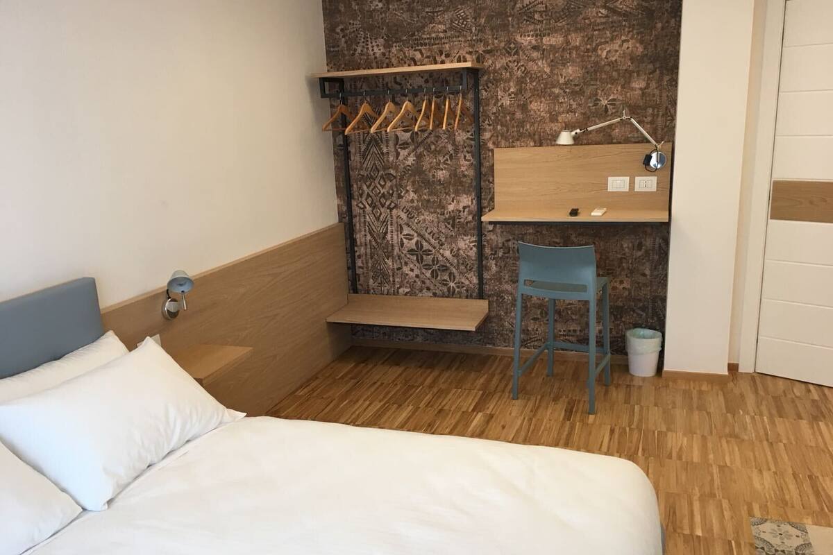 A bedroom with a bed, a desk with a chair, a hanging clothes rack, and a small trash bin.