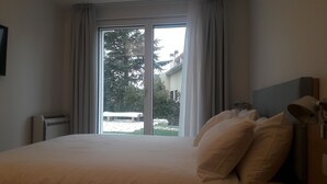 Ein Hotelzimmer mit einem großen Bett, einem Nachttisch, einer Lampe und Blick auf ein Gebäude und Bäume durch eine Schiebetür.