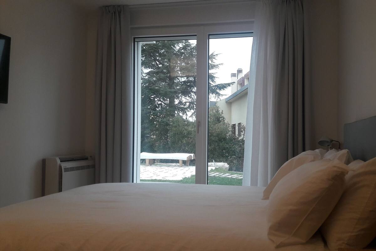 A hotel room with a large bed, a nightstand, a lamp, and a view of a building and trees through a sliding glass door.