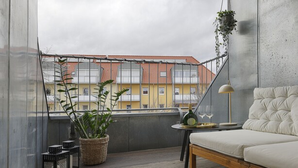 Ein Balkon mit Blick auf Gebäude, ein Blumentopf, ein kleiner Tisch mit Lampe und ein Sofa.