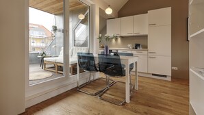 Eine moderne Küche mit weißen Schränken, ein Essbereich mit Glastisch und Stühlen sowie ein Blick ins Freie durch große Fenster.