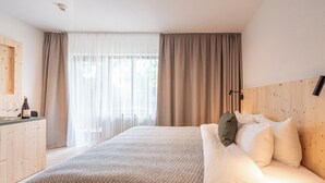 Una habitación de hotel moderna con una cama grande, un cabecero de madera, un escritorio con una botella y vasos, y cortinas de gasa.