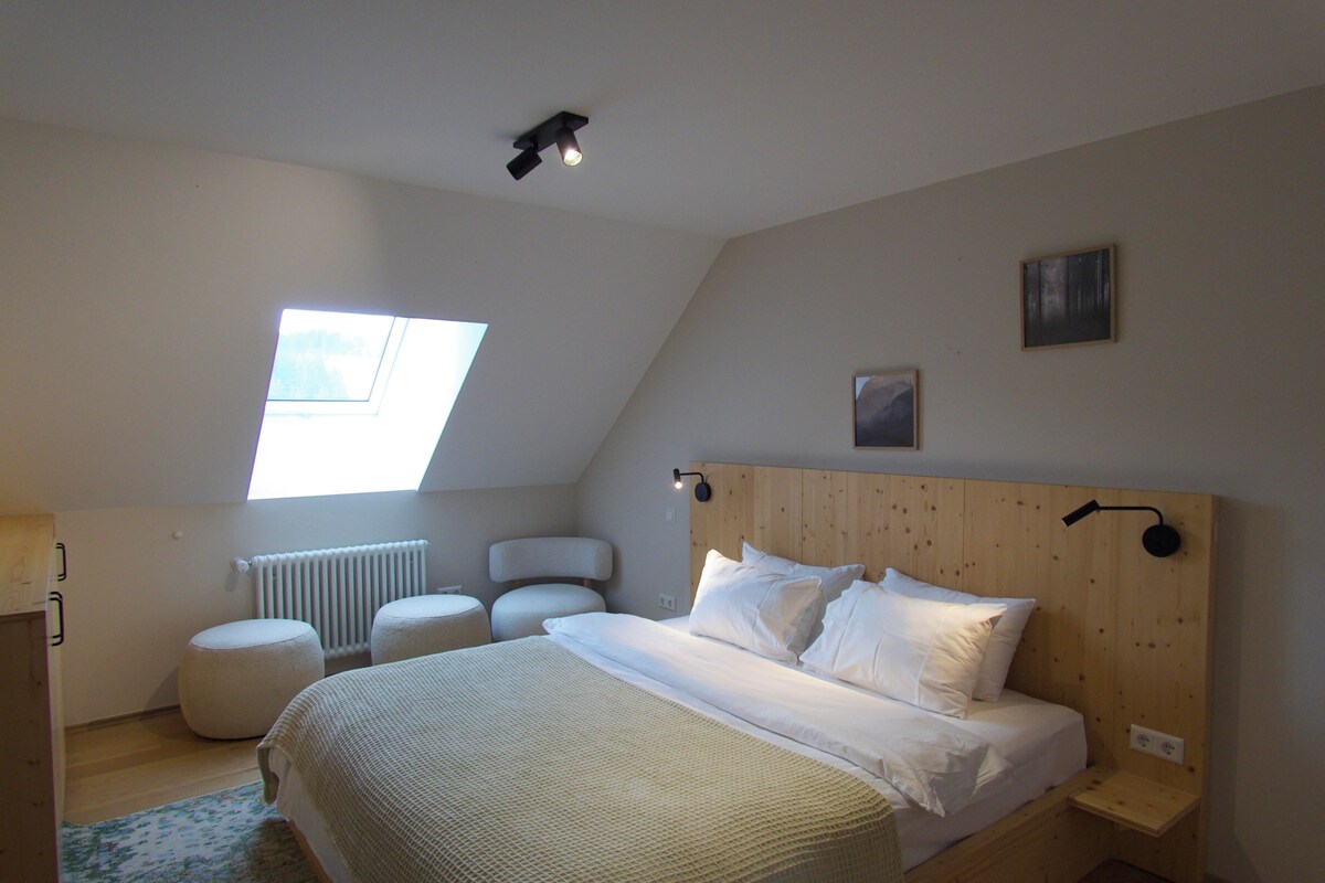 Une chambre avec un plafond en pente, un puits de lumière, un lit avec du linge de maison blanc, une tête de lit en bois et une petite table de chevet.