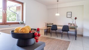 Una cocina moderna con un bol de limones y tomates en la encimera, una zona de comedor con una mesa y sillas, y un ventanal con vistas a la vegetación.