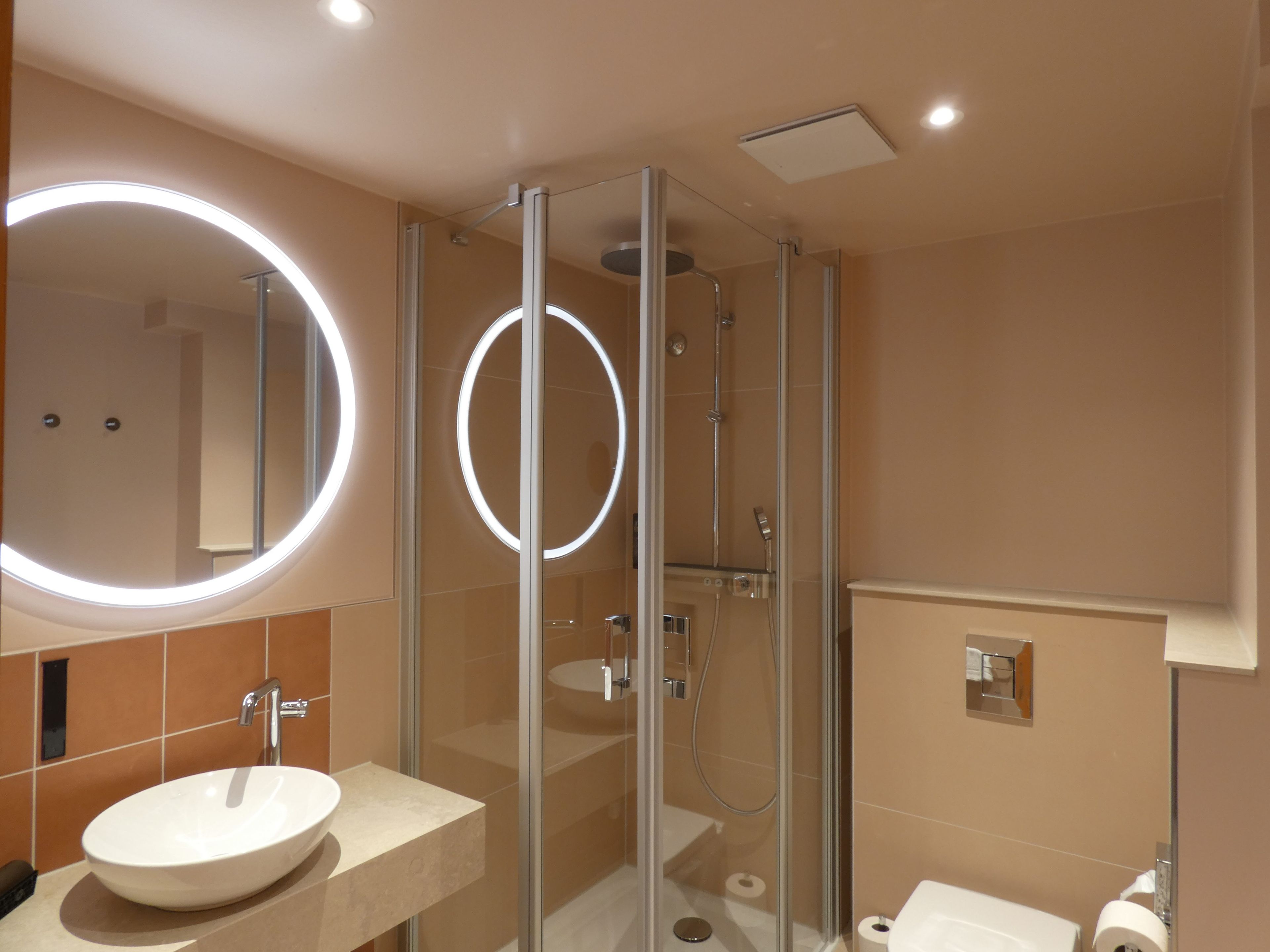 Une salle de bain moderne avec une douche à parois de verre, un grand miroir rond doté de lumières et un lavabo blanc.