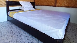 Una cama individual con cabecera de madera, ropa de cama blanca y una manta amarilla. El cuarto tiene piso de baldosas y paredes cubiertas de alfombras tejidas.