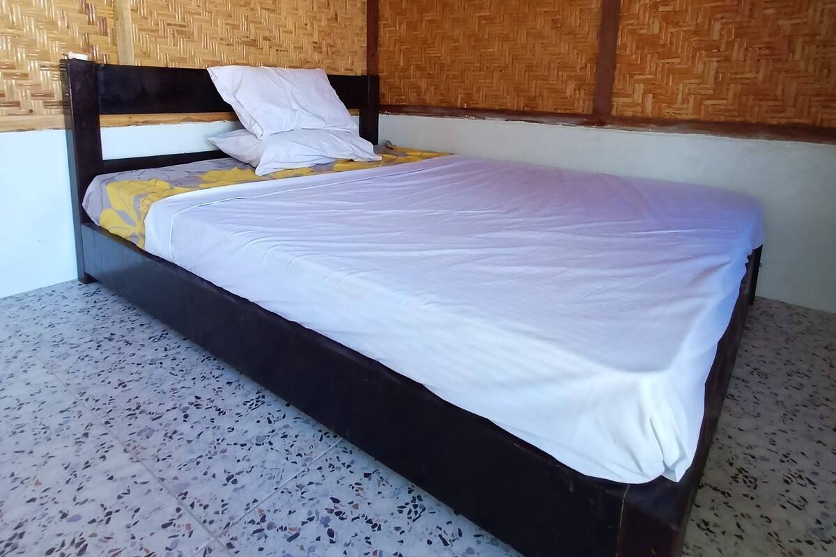 A single bed with a wooden headboard, white bedding, and a yellow blanket. The room has a tiled floor and walls covered with woven mats.