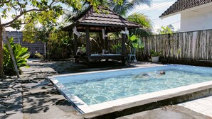 Una piscina con borde de baldosas, rodeada por un jardín con diversas plantas y una estructura de pérgola.