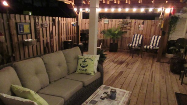 An outdoor patio with a wooden deck, a cushioned sofa with anchor-themed pillows, striped chairs, and a small table with a drink. - THE " BOAT ANCHOR LOUNGE"  UPSCALE LUXURY (North Augusta)