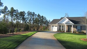 A residential house with a driveway leading to a garage, surrounded by a well-maintained lawn and tall trees. - THE " BOAT ANCHOR LOUNGE"  UPSCALE LUXURY (North Augusta)