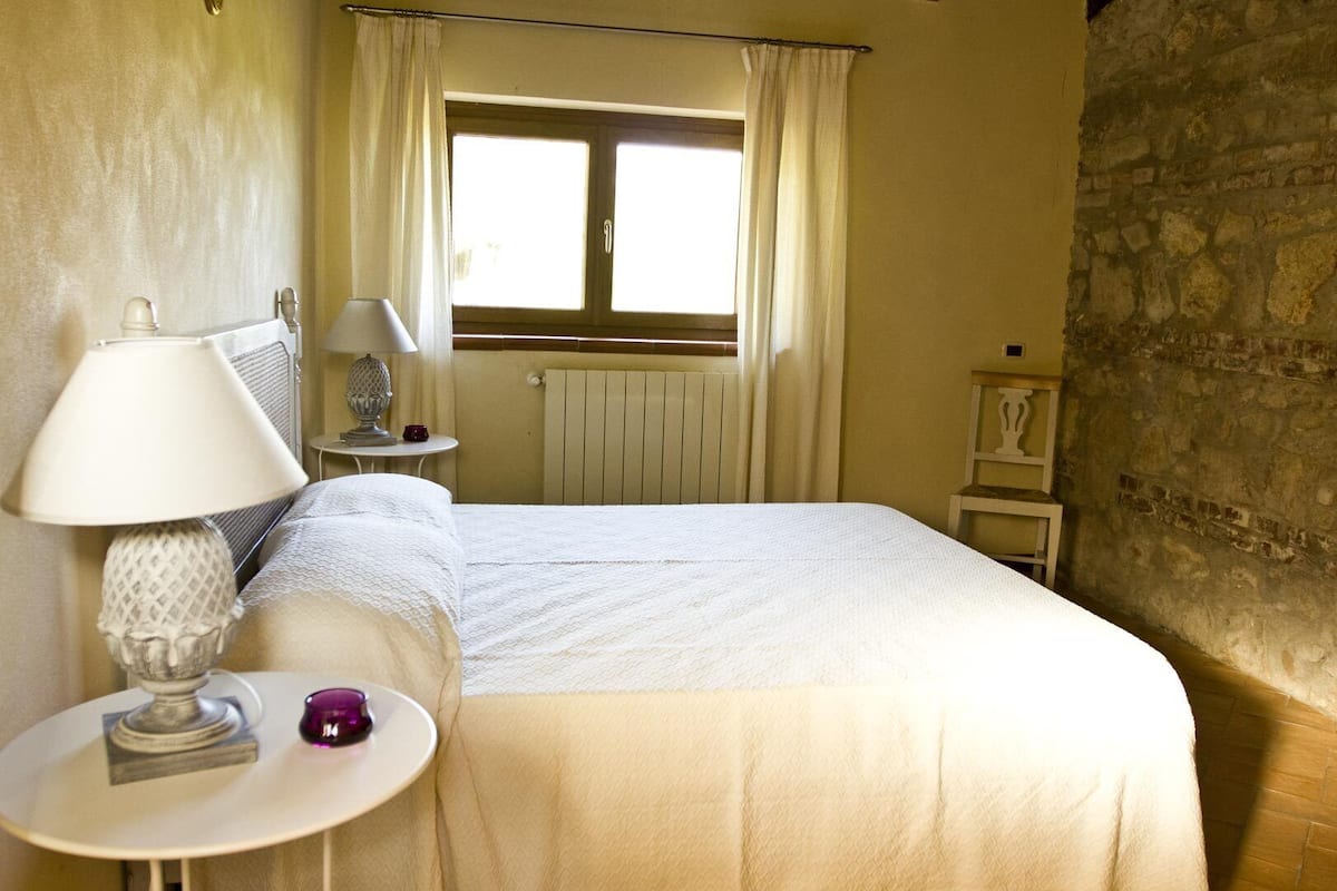 Ein Hotelzimmer mit einem Bett, einem Nachttisch mit Lampe, einem kleinen Tisch mit Dekoration, einem Fenster mit Vorhängen und einer Steinwand.