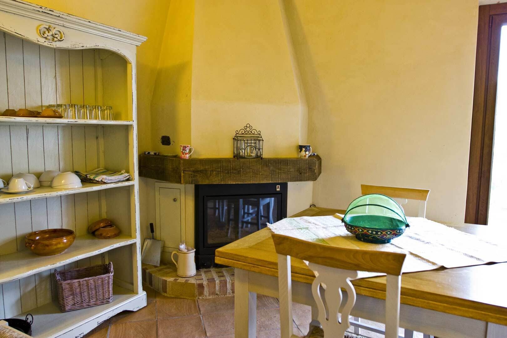 Una habitación acogedora con chimenea, estantes de madera con vajilla, una mesa con un tazón verde y una cesta de mimbre.