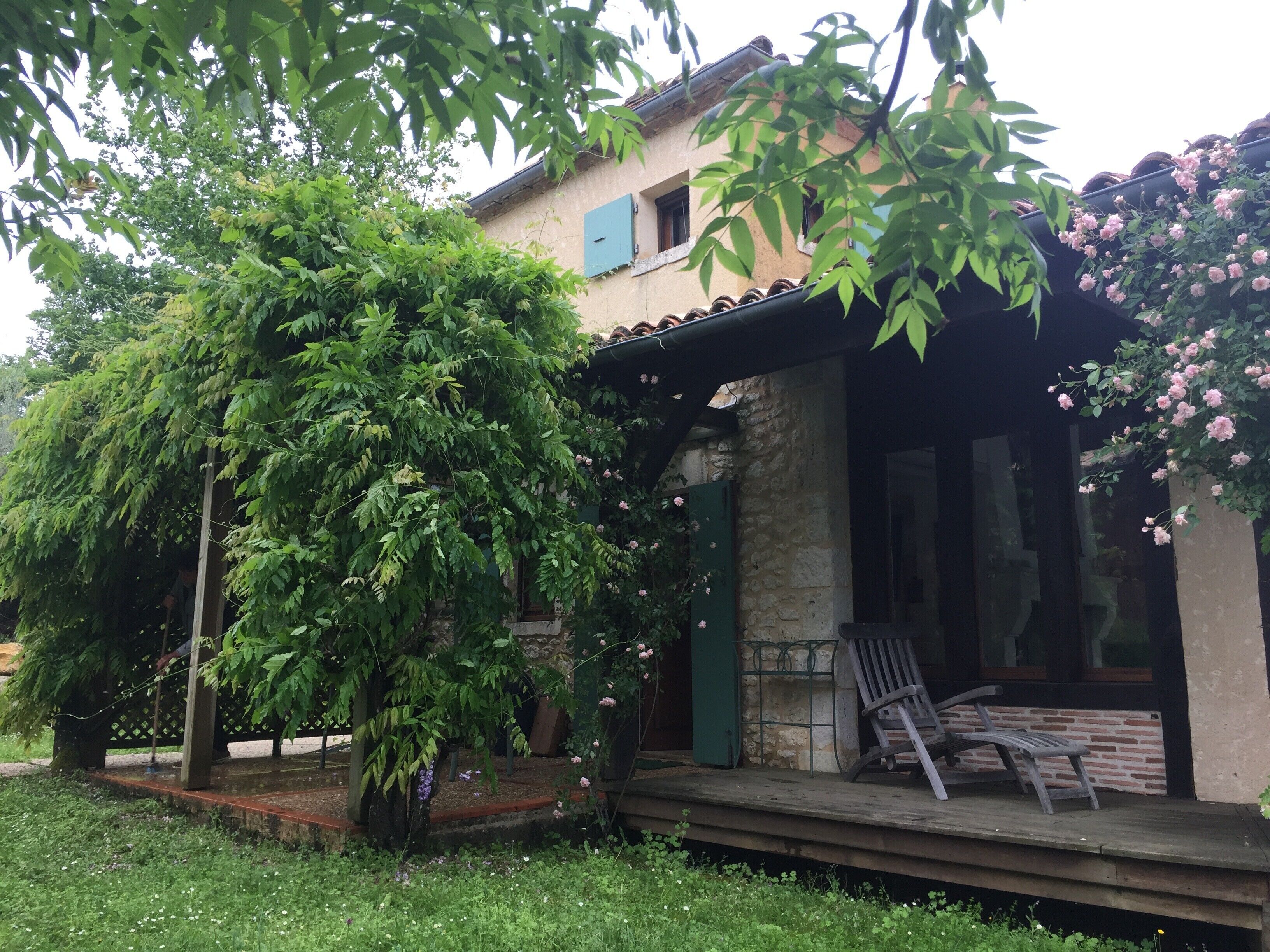 A rustic house with a wooden porch, a stone wall, and a garden with a large tree and blooming flowers.