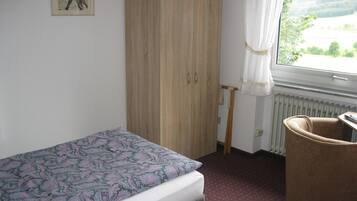 A bedroom with a bed, a wooden wardrobe, a chair, a radiator, and a window with a view of greenery.