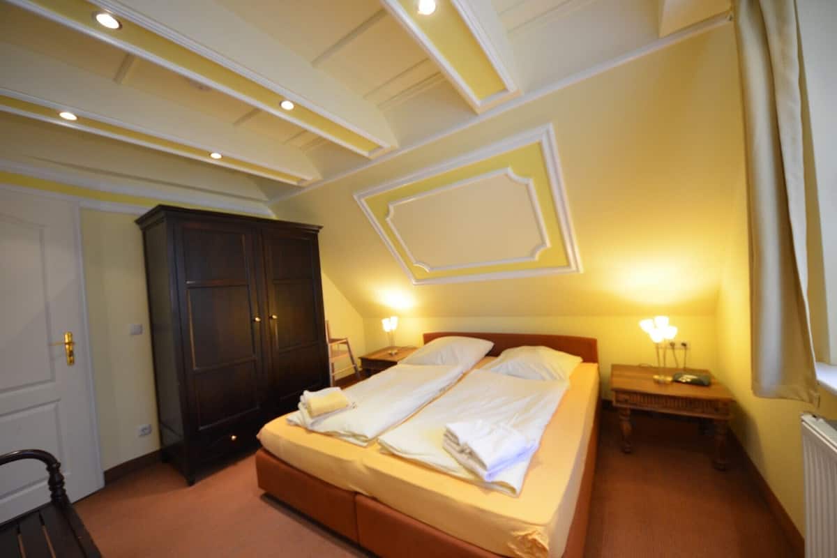 A hotel room with a double bed, two bedside tables with lamps, a dark wooden wardrobe, and a white door.