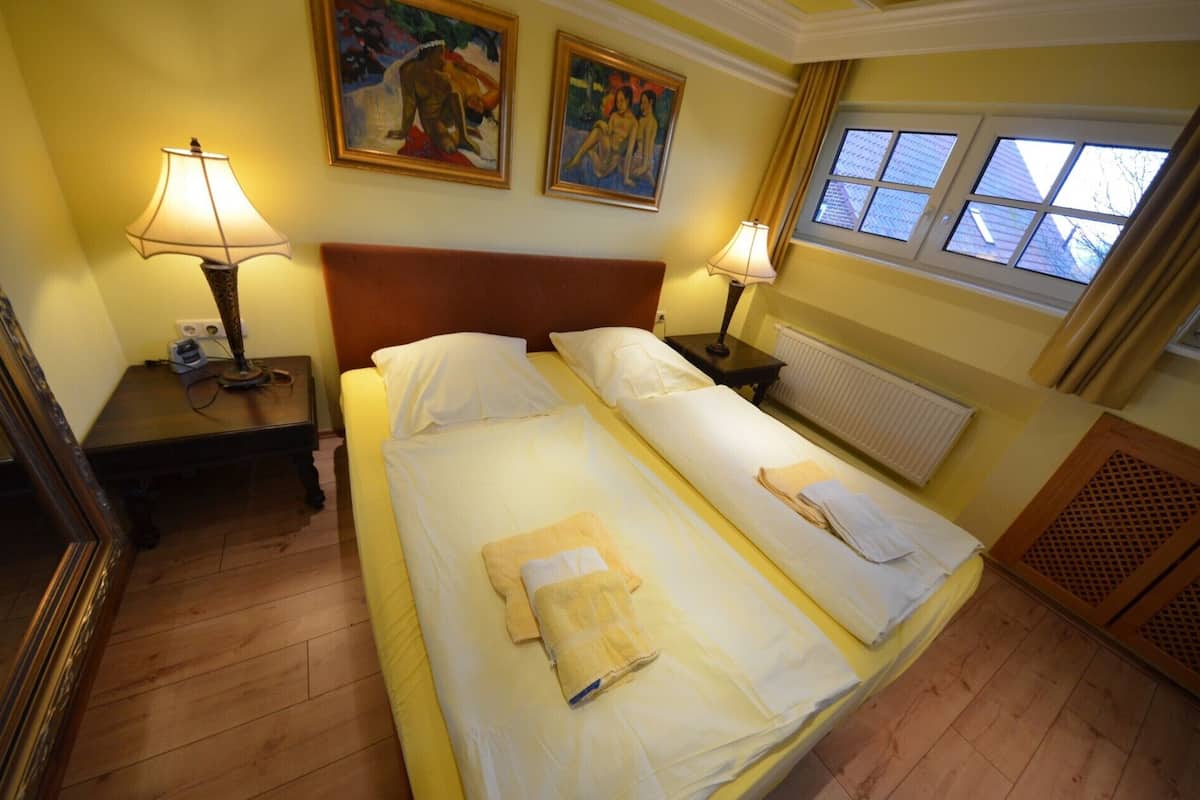 A double bed with two pillows and a folded towel, a nightstand with a lamp, a painting on the wall, and a window with curtains.