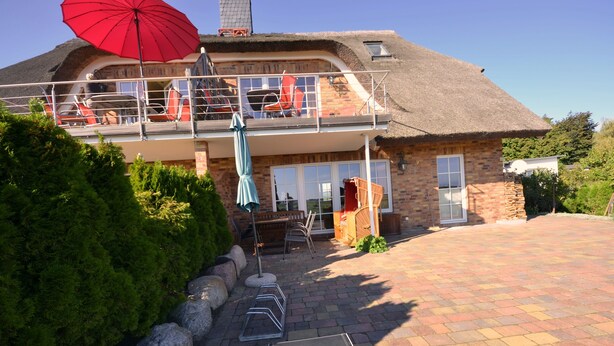 Een tweelaags huis met een balkon, een rode parasol en een geplaveid pad.