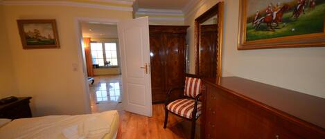 A hotel room with a wooden floor, a bed with a white bedspread, a chair, a wooden dresser, and a painting on the wall.