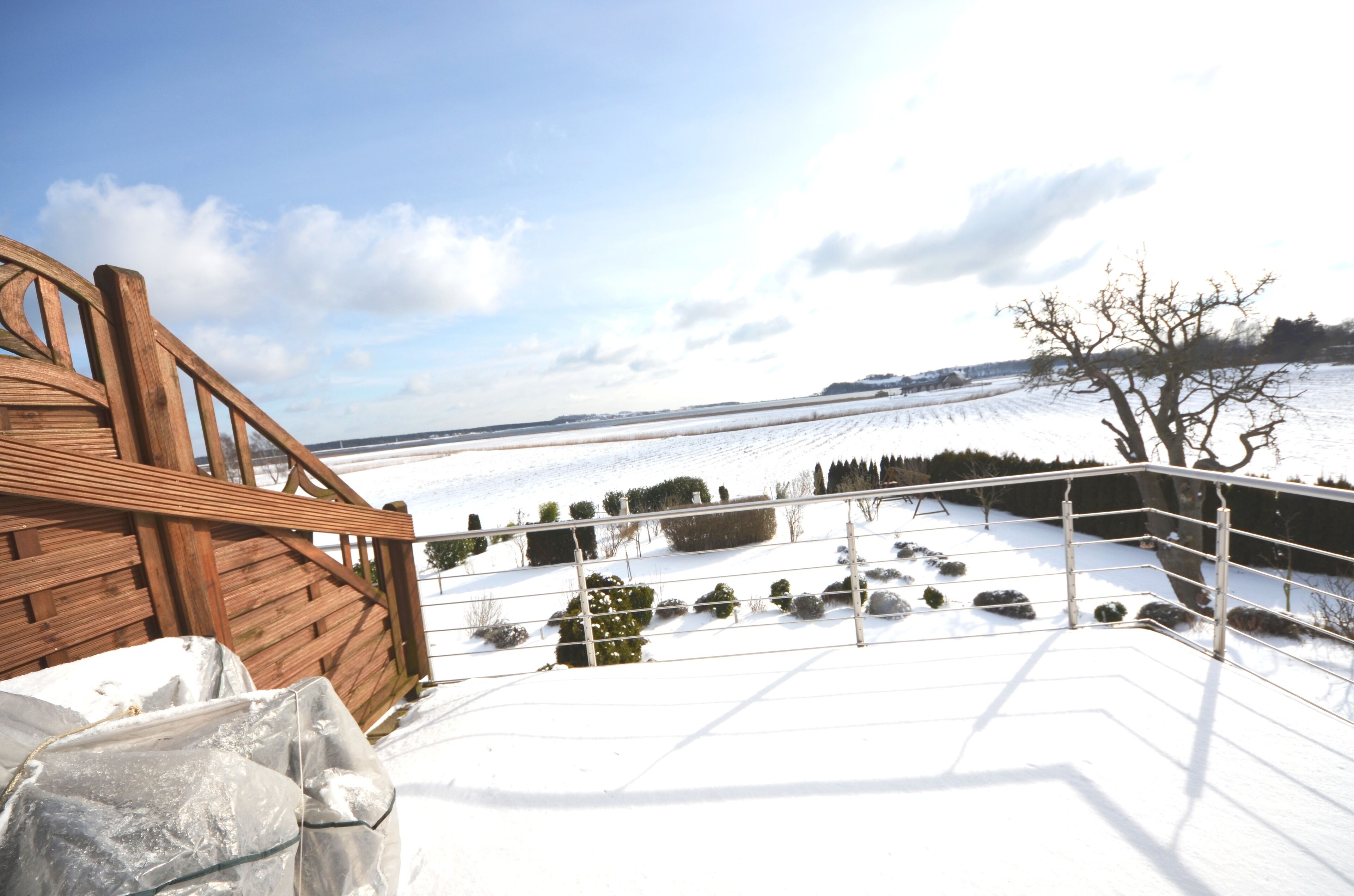 Una terraza cubierta de nieve con baranda de madera, cerca metálica y vista a un paisaje nevado con árboles y una colina distante.