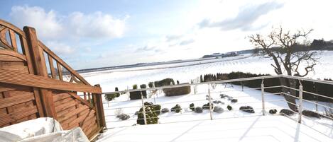 A snow-covered deck with a wooden railing, a metal fence, and a view of a snowy landscape with trees and a distant hill.