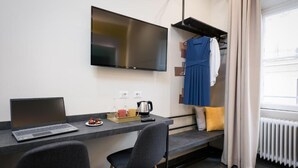 Une chambre d’hôtel moderne équipée d’un téléviseur à écran plat fixé au mur, d’un bureau avec un ordinateur portable, d’une petite table basse et d’un coin salon avec un banc.