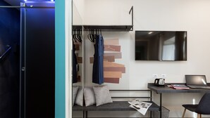 Une chambre d’hôtel compacte comprenant un lit, un bureau avec un ordinateur portable, une télévision et un coin salon avec un banc et des coussins.