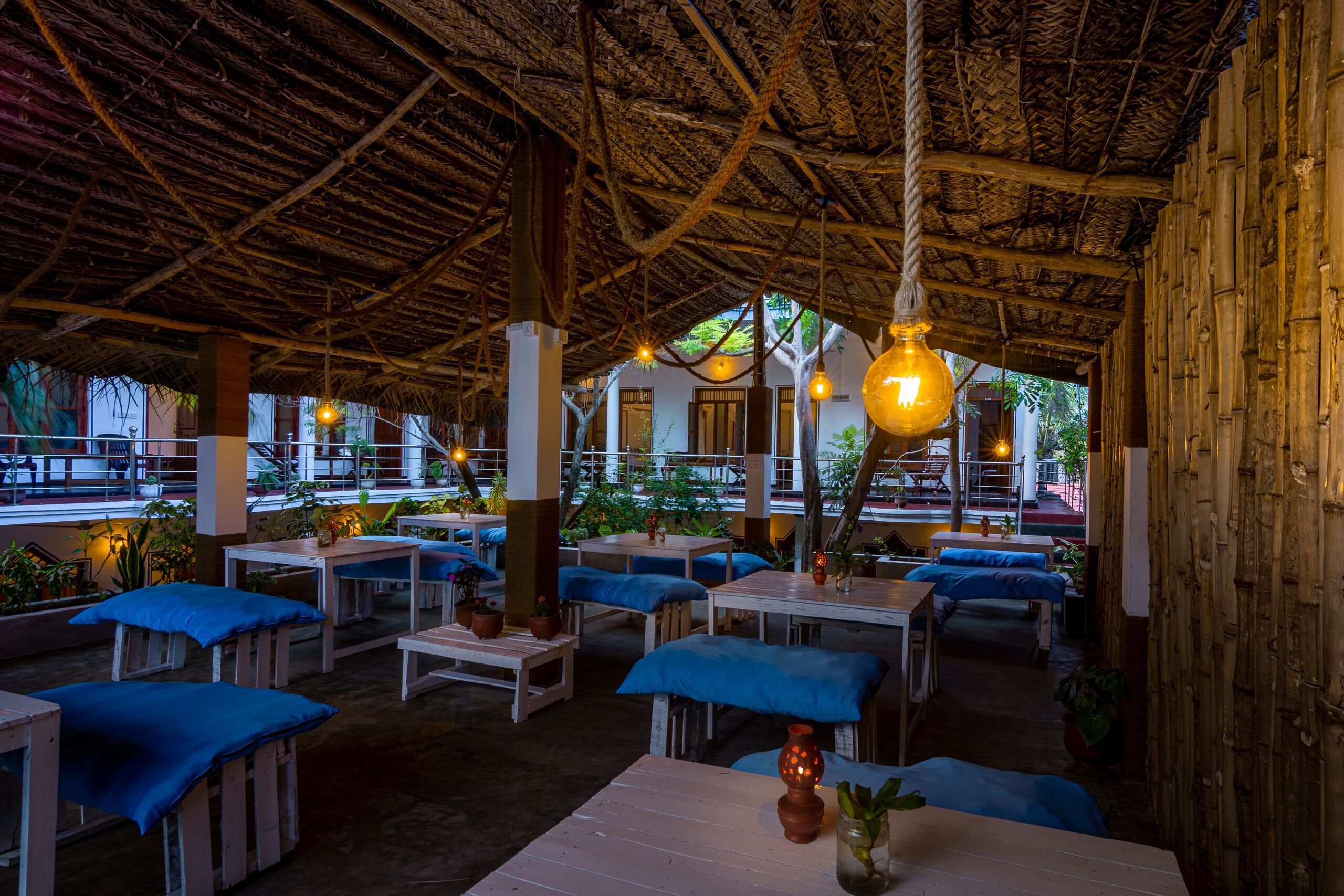 Un área de comedor al aire libre con mesas y bancos de madera, techo de paja, luces colgantes y plantas en macetas.