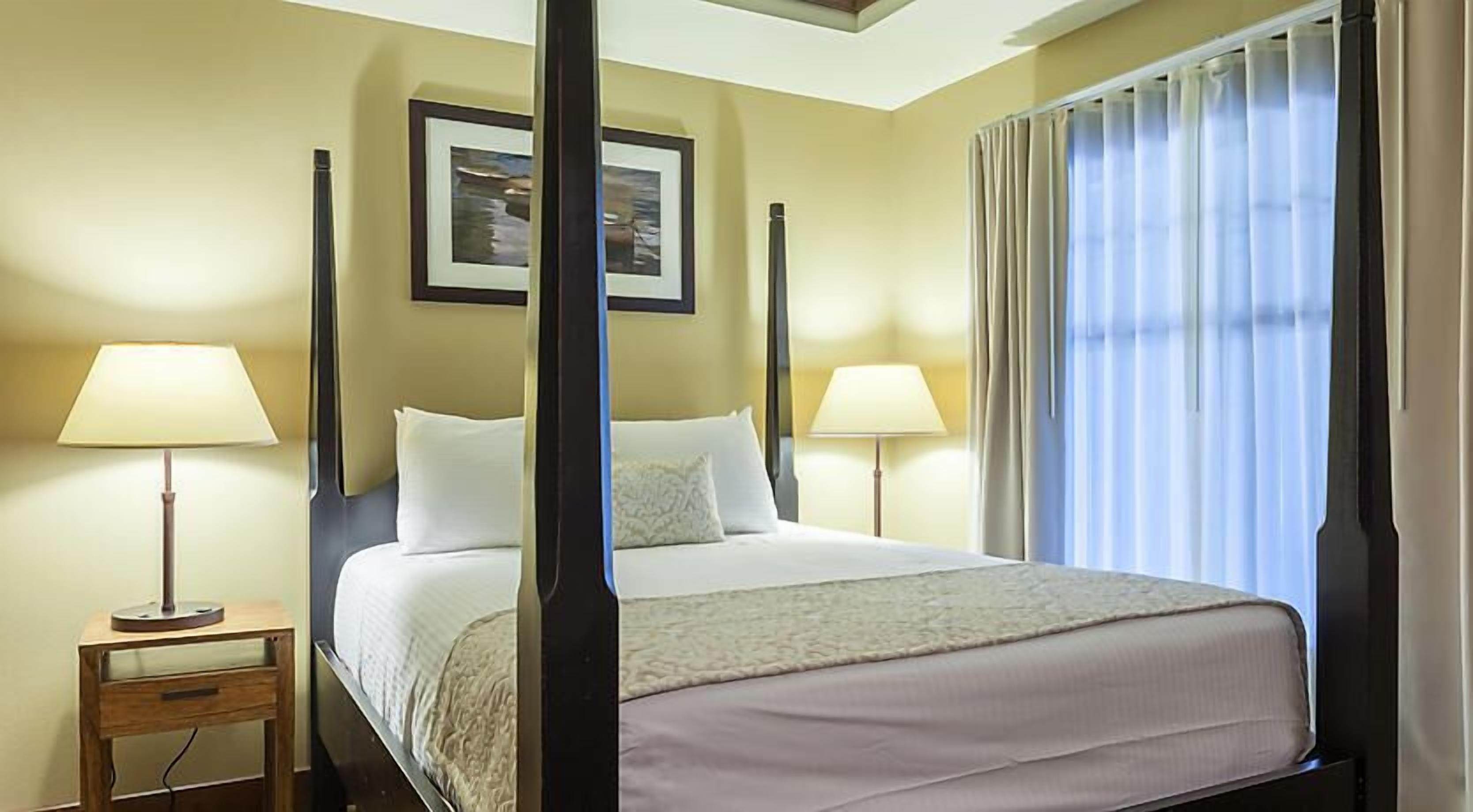 A four-poster bed with a white bedspread, two bedside lamps, a framed picture on the wall, and a window with curtains.
