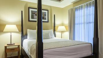 A four-poster bed with a white bedspread, two bedside lamps, a framed picture on the wall, and a window with curtains.
