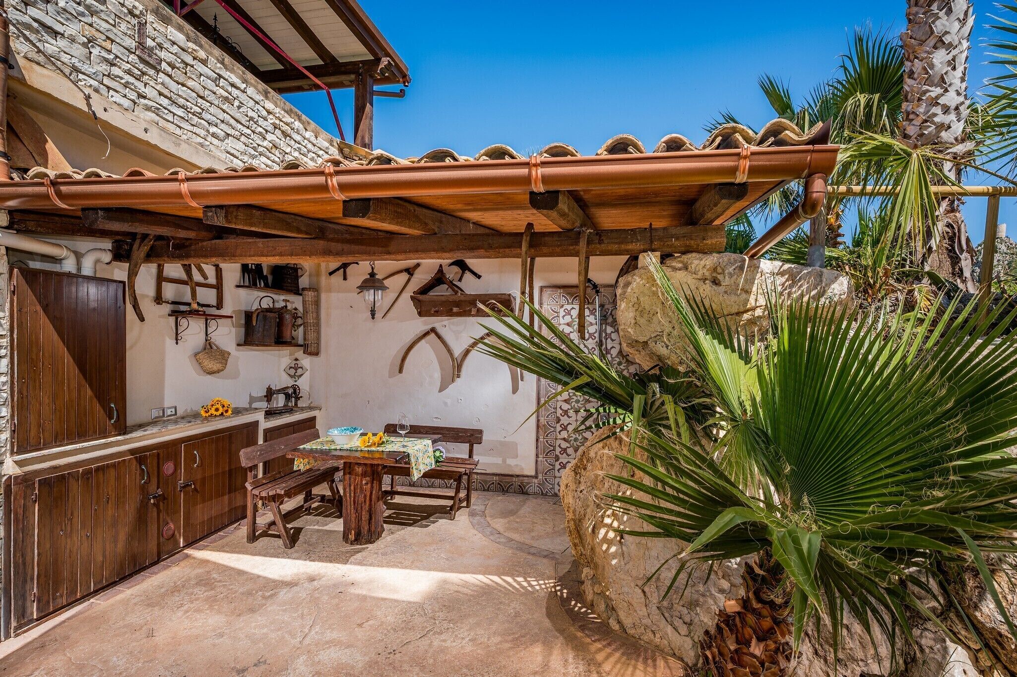 Un espacio de comedor al aire libre con estilo rústico, una pared de piedra, muebles de madera y una palmera.