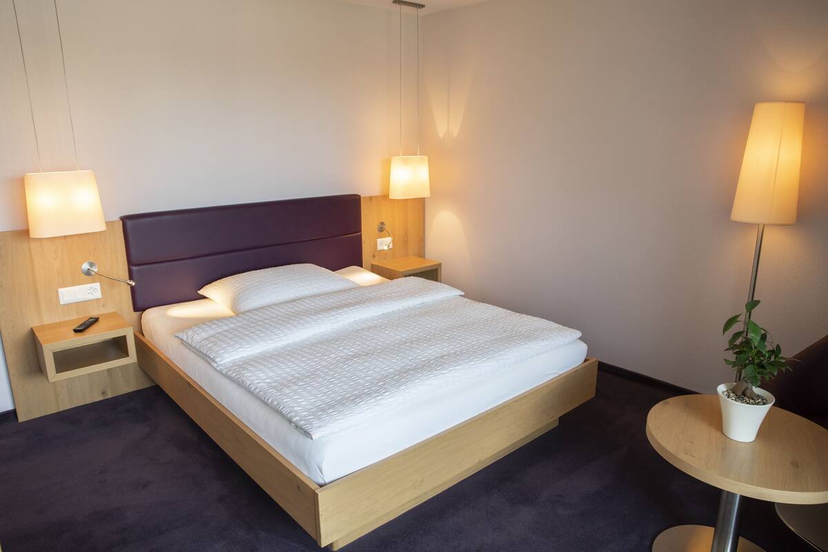 A neatly made bed with a wooden headboard, two bedside tables with lamps, a small round table with a plant, and a wall socket.