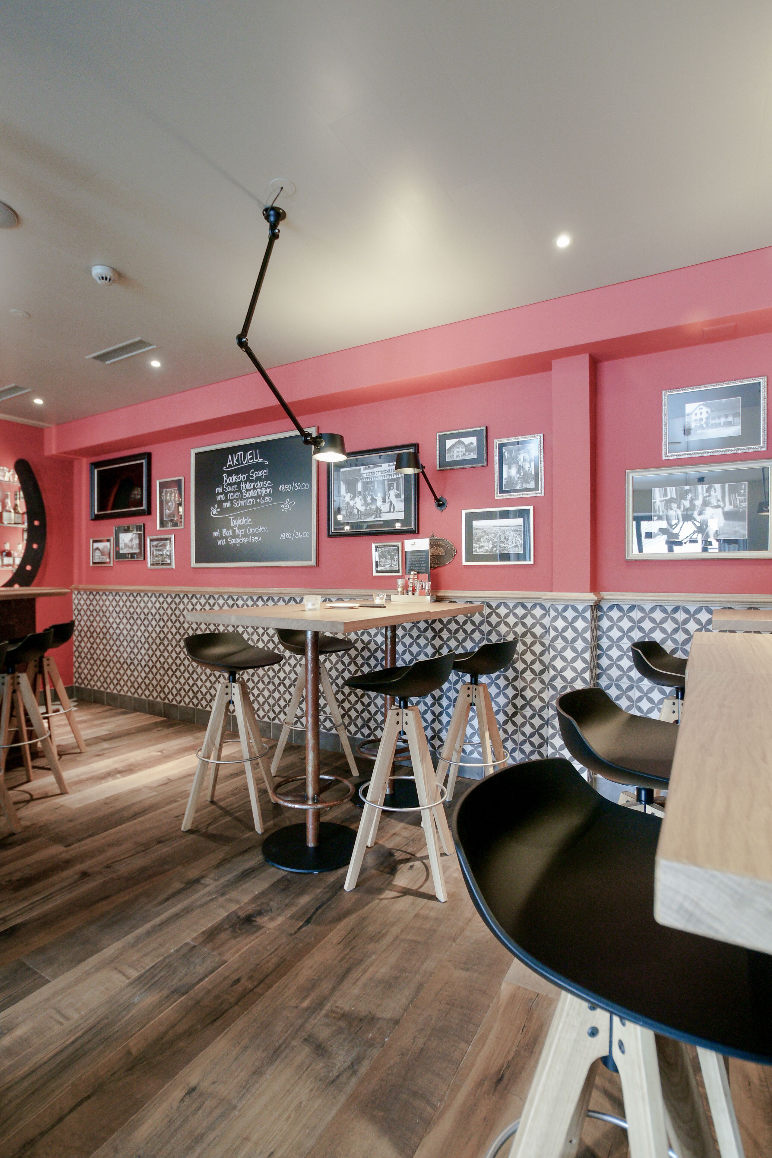 A restaurant interior with a red wall, black and white patterned backsplash, wooden flooring, and black bar stools.